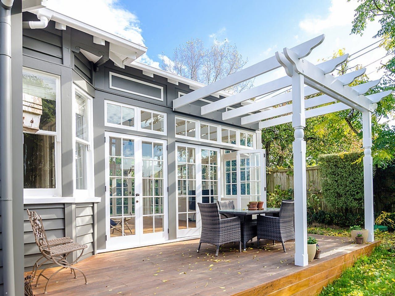 A wooden deck with patio furniture and a white pergola, attached to a gray house with white-framed French doors.