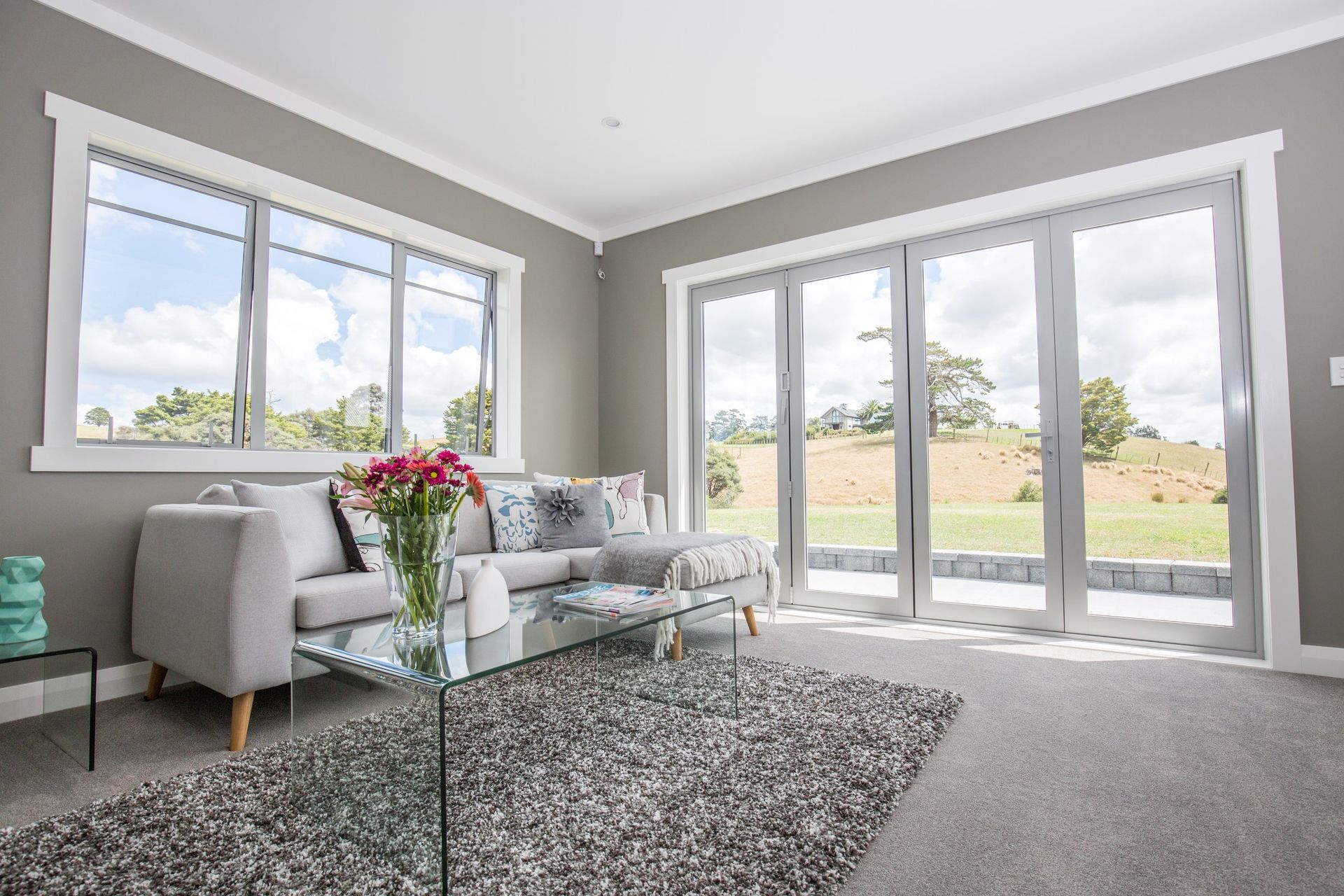 A bright living room with a grey sofa, glass coffee table, and large windows looking out onto a grassy landscape.