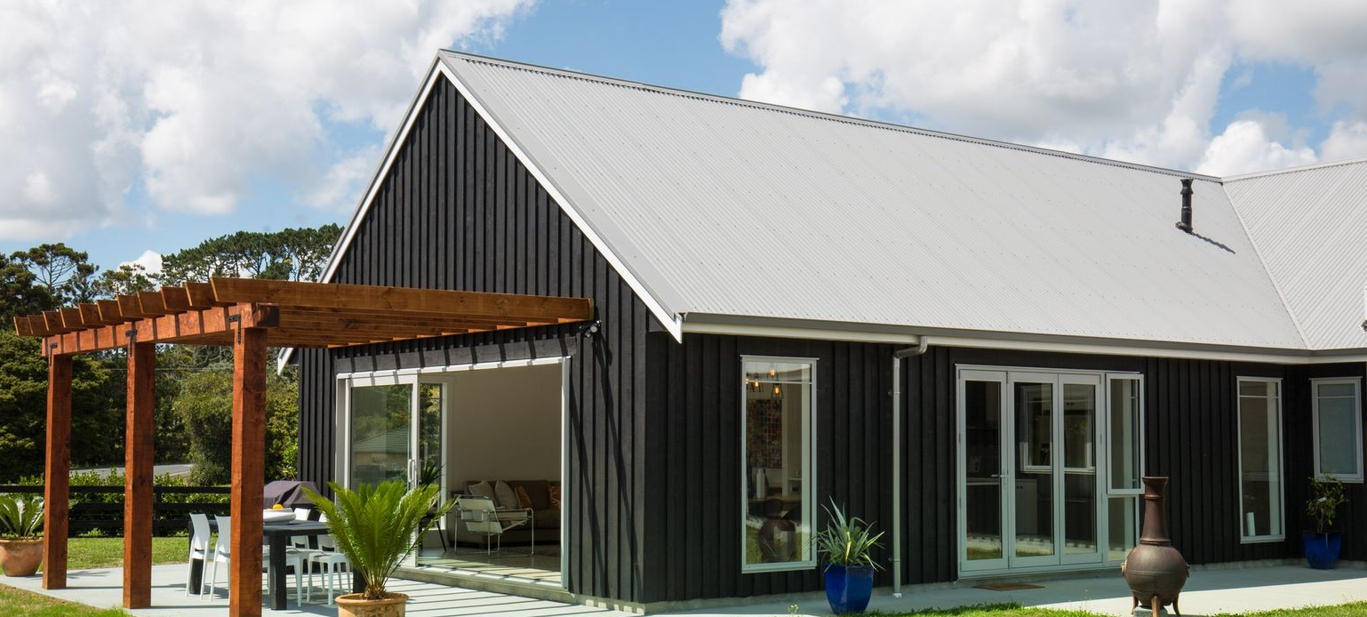A black house with a white roof and wooden pergola, featuring open sliding doors leading to a patio with outdoor furniture.