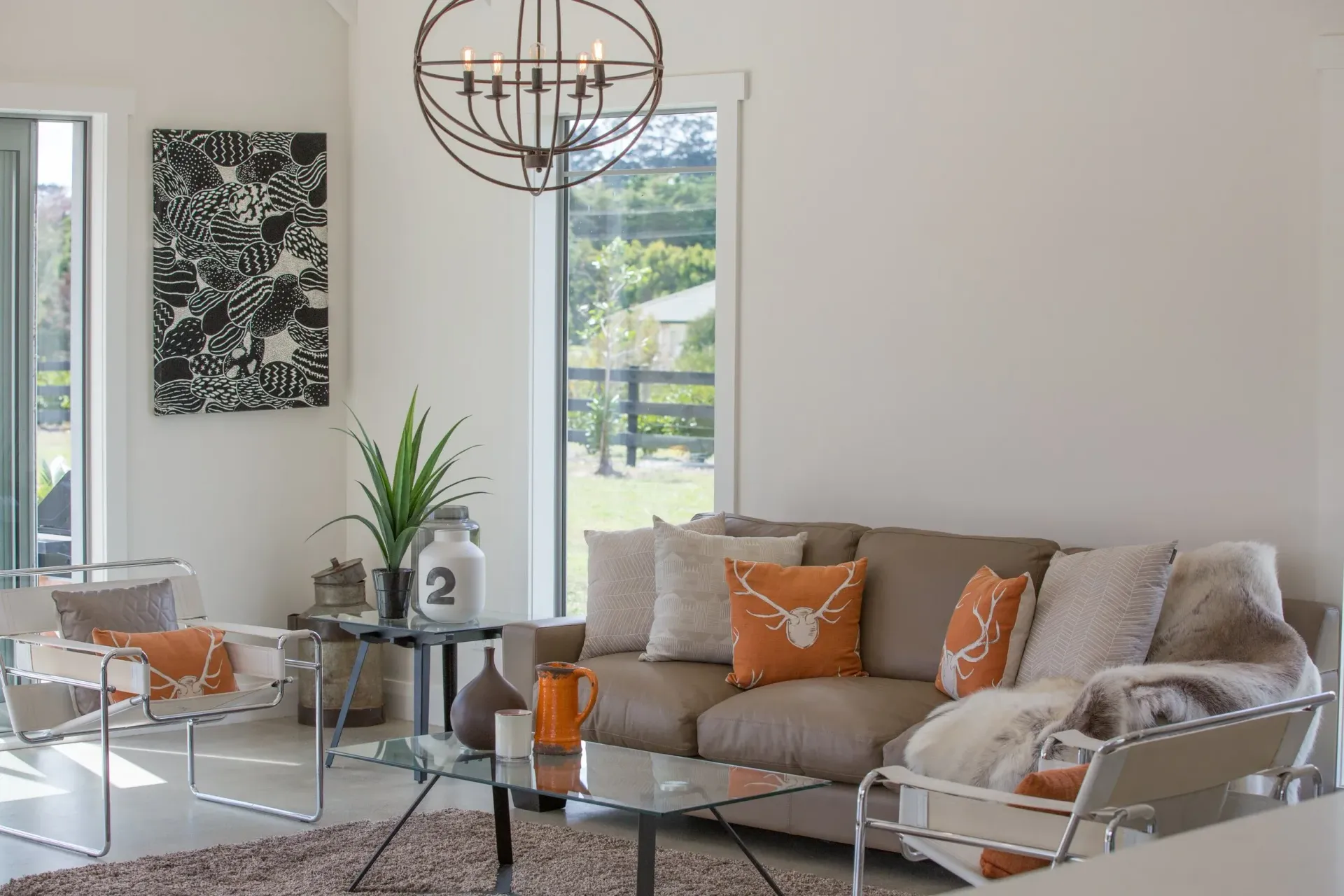 A modern living room with a beige sofa featuring orange cushions, a glass coffee table, and a black-and-white abstract art.