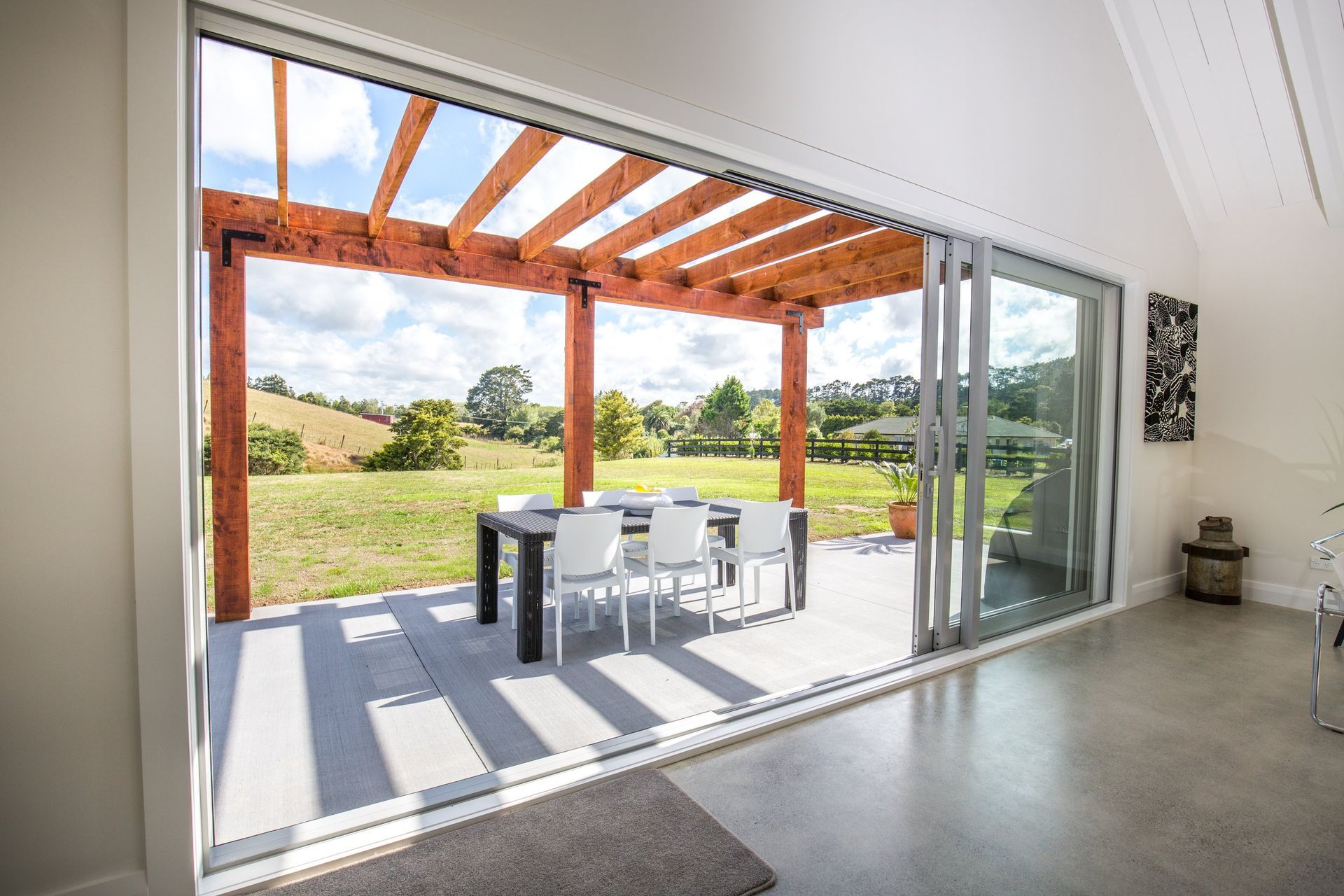 A spacious patio with a wooden pergola and dining set, viewed through large sliding glass doors from an indoor room.