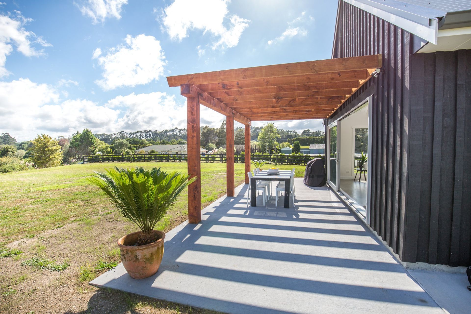 A wooden pergola over a concrete patio with a dining set and potted plant, attached to a dark-sided house on a sunny day.