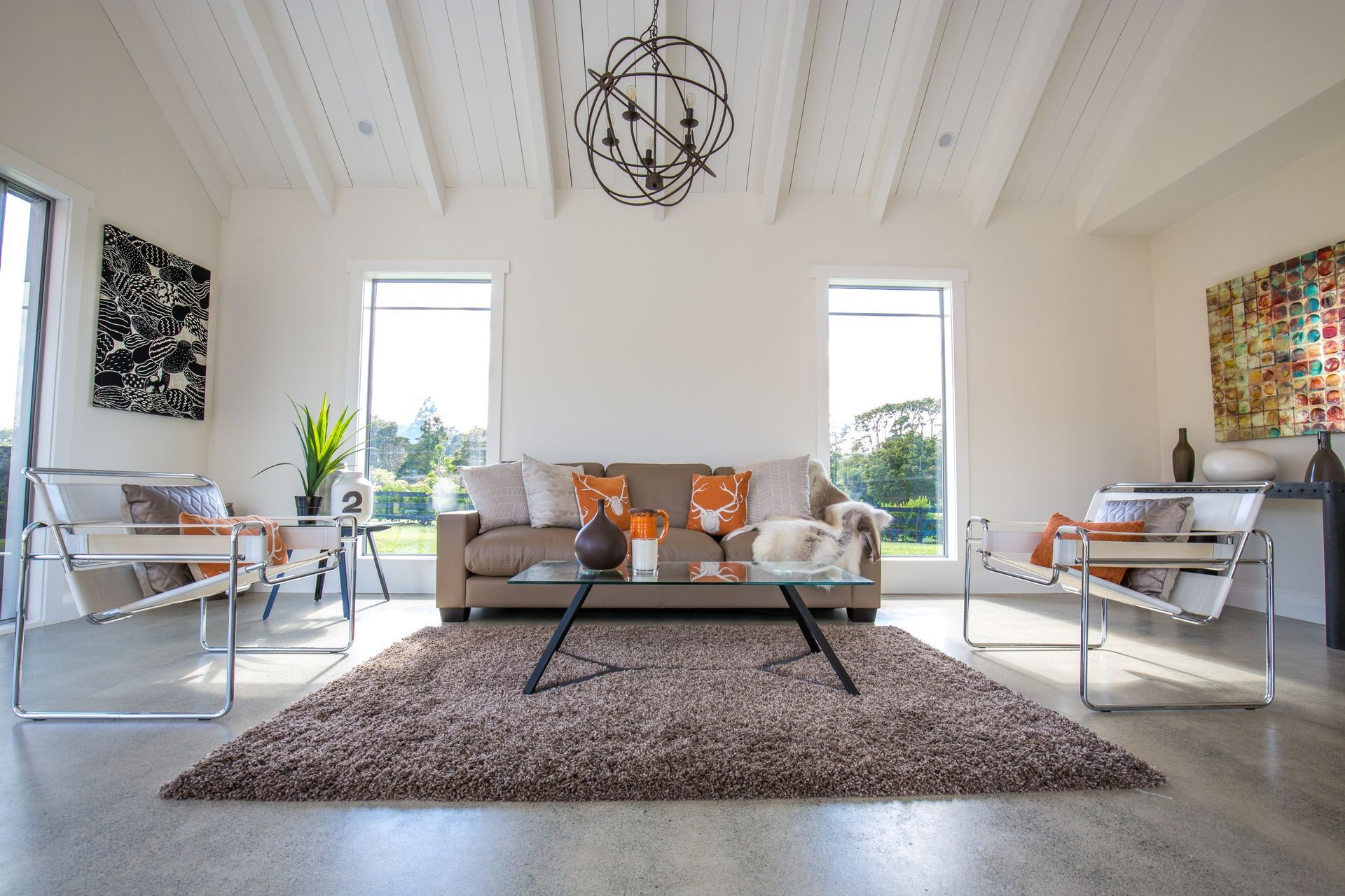 A modern living room with a beige sofa, two white lounge chairs, a brown rug, and windows under a vaulted ceiling.