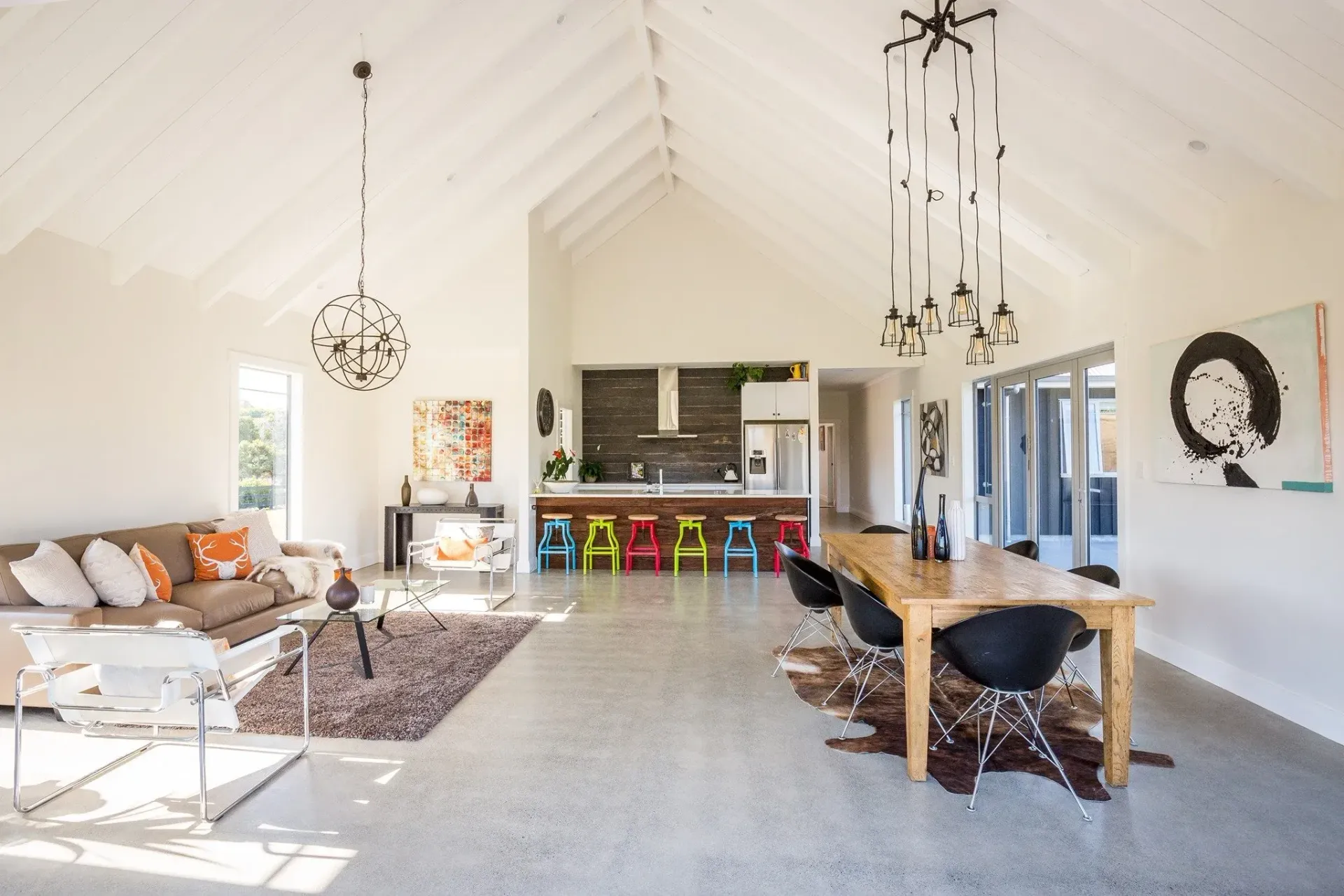 A bright, open-plan living and dining area with vaulted ceilings, white walls, colorful stools, and modern chandeliers.