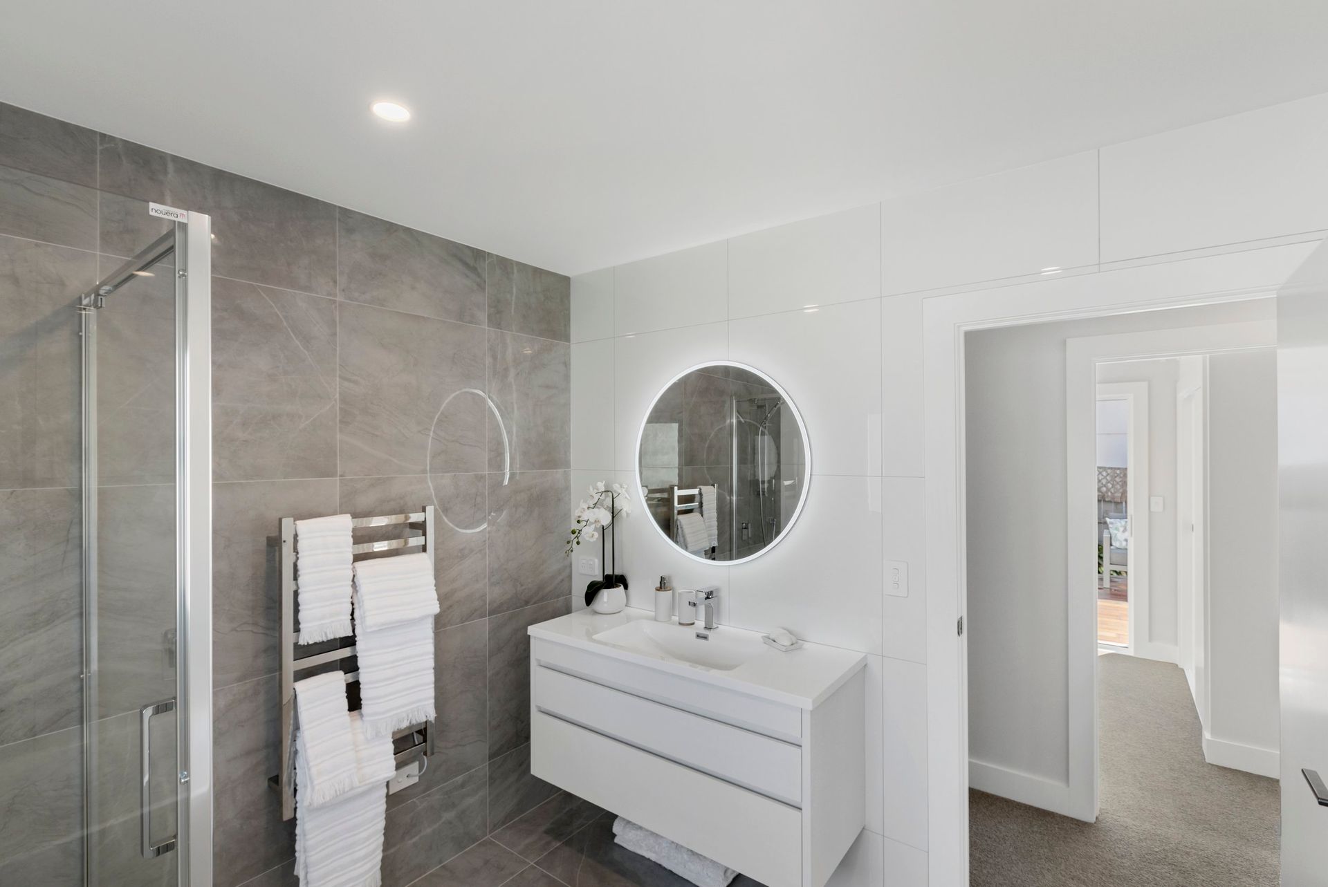 A modern bathroom featuring grey tiled walls, a glass walk-in shower, a white floating vanity, and a backlit round mirror.