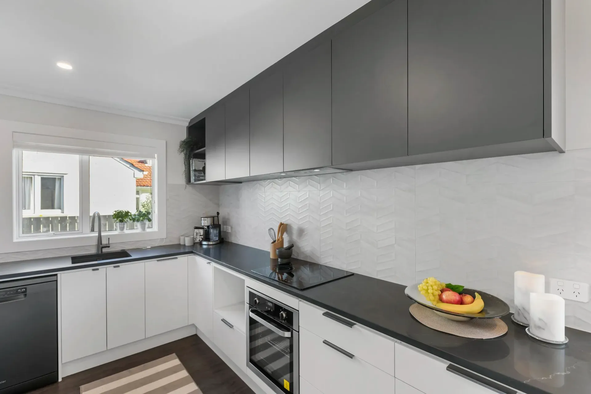 A modern kitchen featuring white lower cabinets, charcoal upper cabinets, a dark countertop, and a window above the sink.