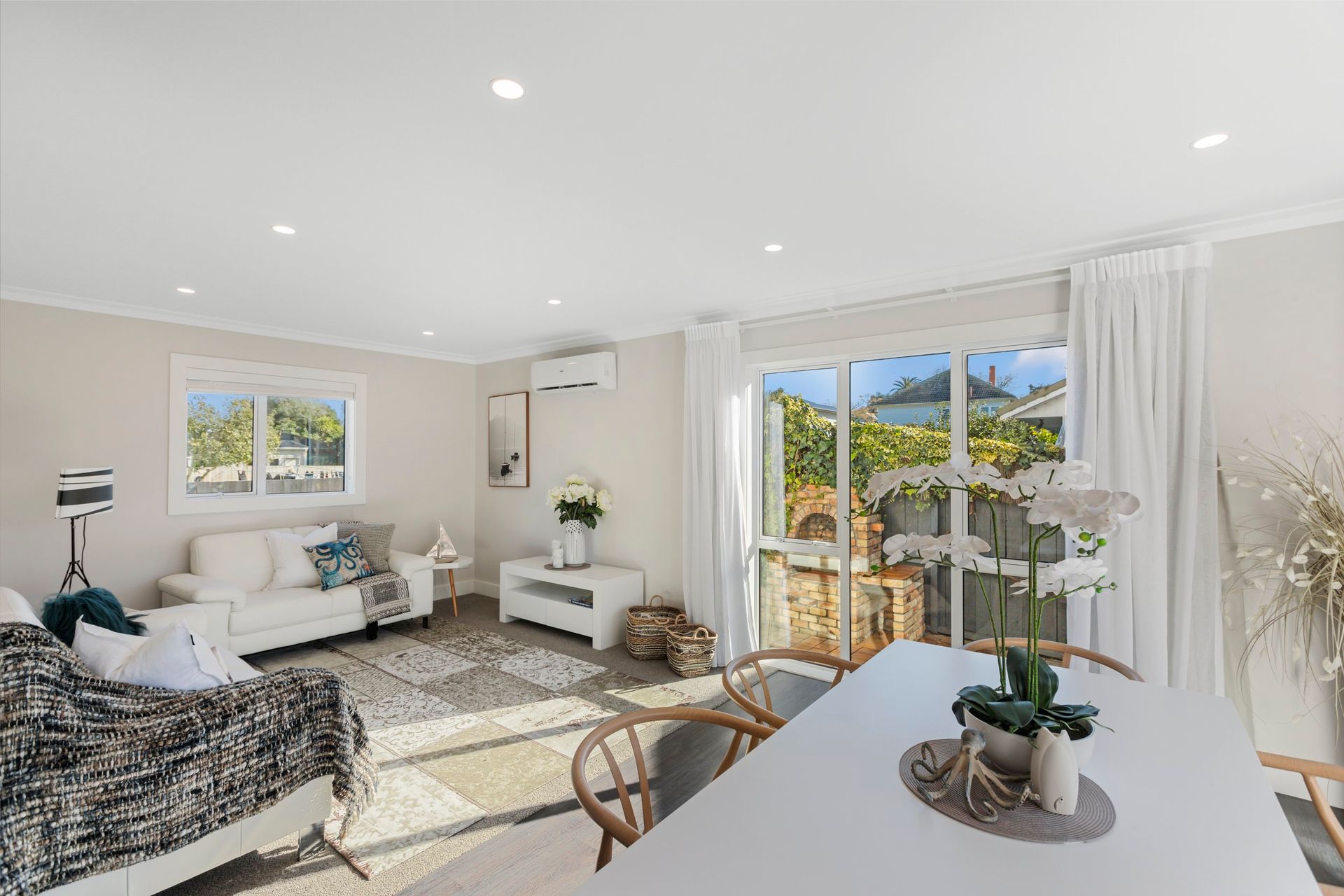 A bright, open-plan living and dining area with white sofas, a patterned rug, a dining table, and large sliding doors.