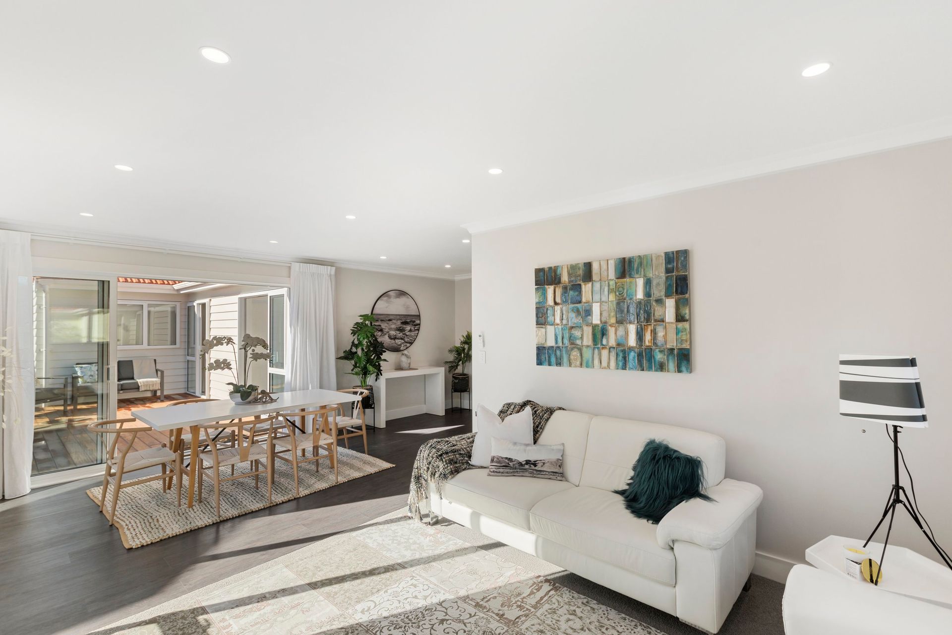 A bright living and dining room with a white sofa, a long dining table, dark wood floors, and a blue abstract painting.