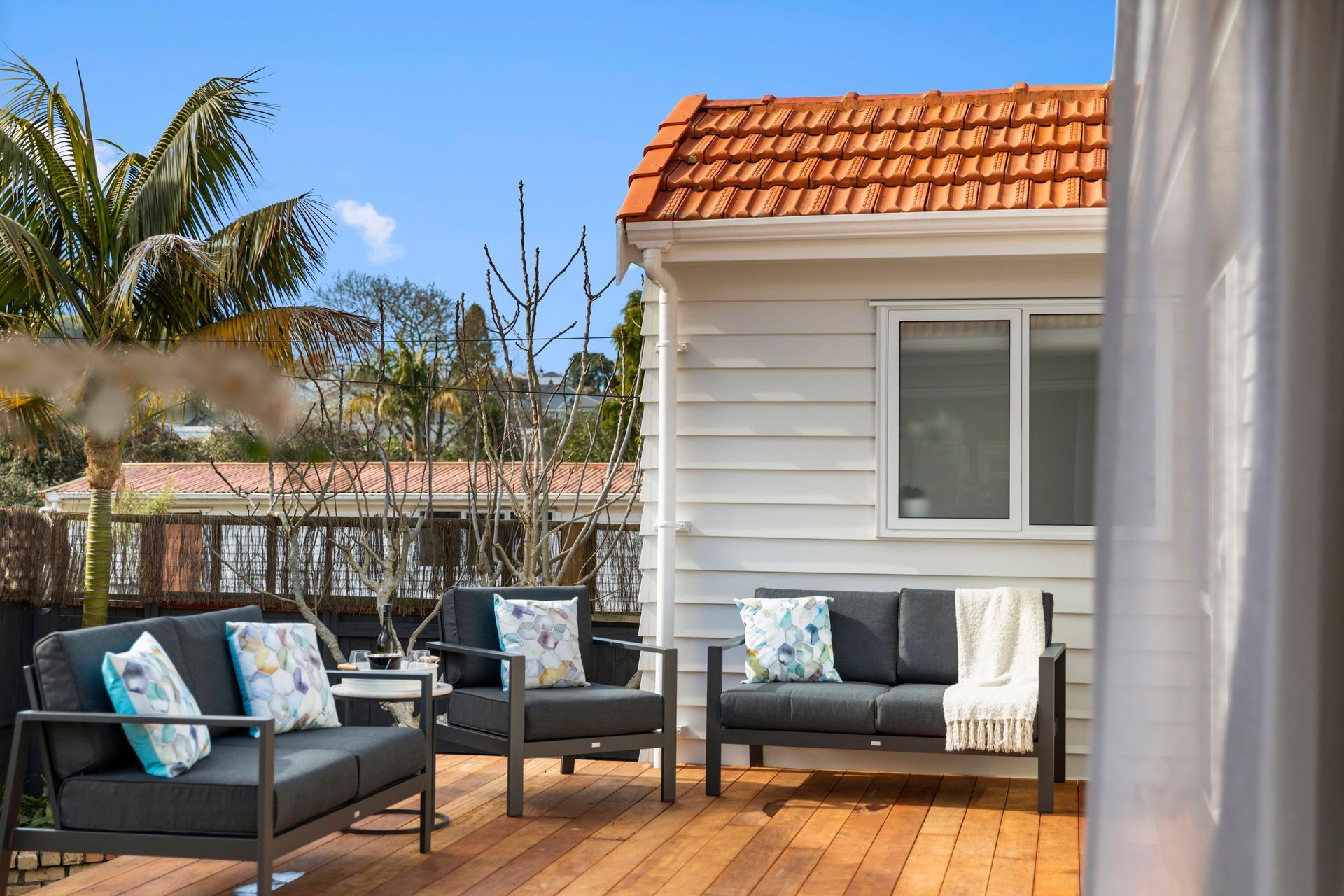 Outdoor patio with two gray cushioned couches and accent pillows on a wooden deck next to a small white building.