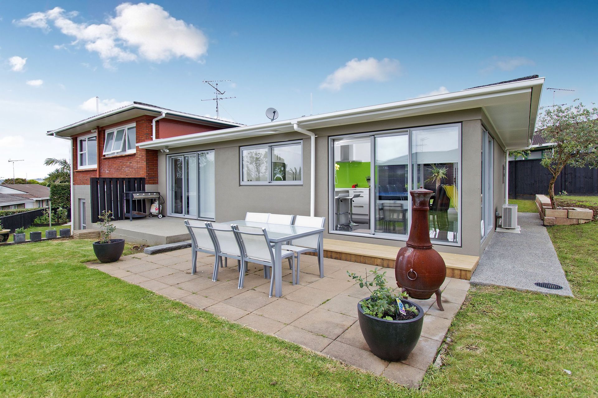 A single-story house with a patio, outdoor dining set, and potted plants on a lawn under a partly cloudy sky.