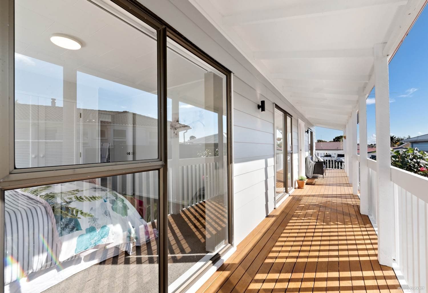 A long, wooden covered deck with white railings and walls, featuring sliding glass doors reflecting an interior room.