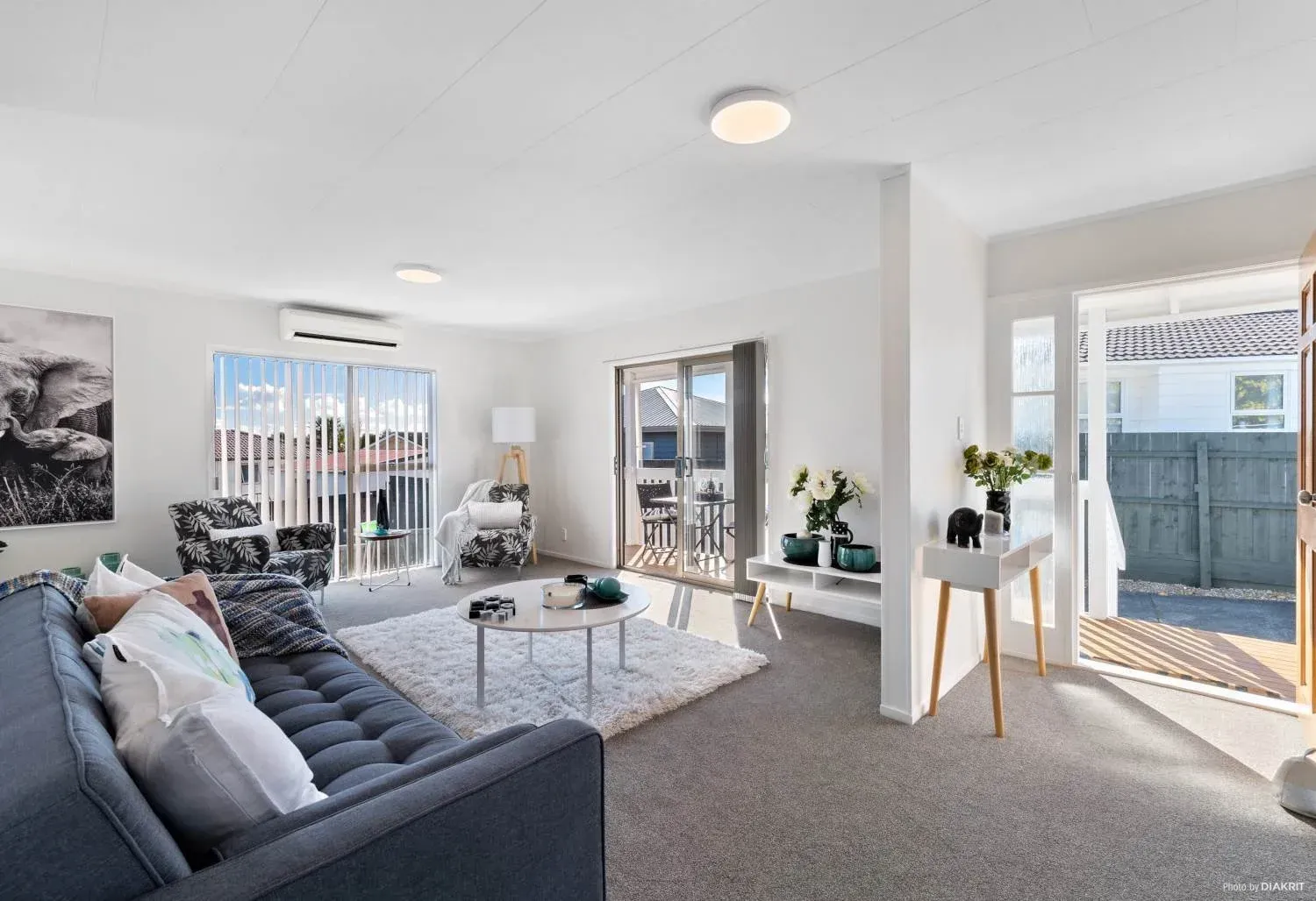 A bright, modern living room with a gray sofa, a circular coffee table, light carpeting, and a doorway to a patio area.