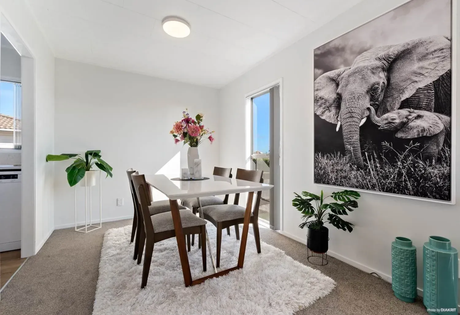 A dining area features a white table with four chairs on a white rug, a large elephant print, and green plants.
