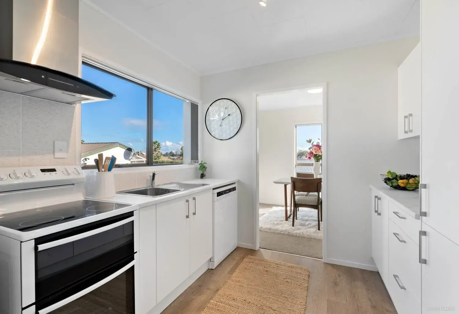 A modern, minimalist kitchen with white cabinets, stainless steel appliances, and a window overlooking a sunny sky.