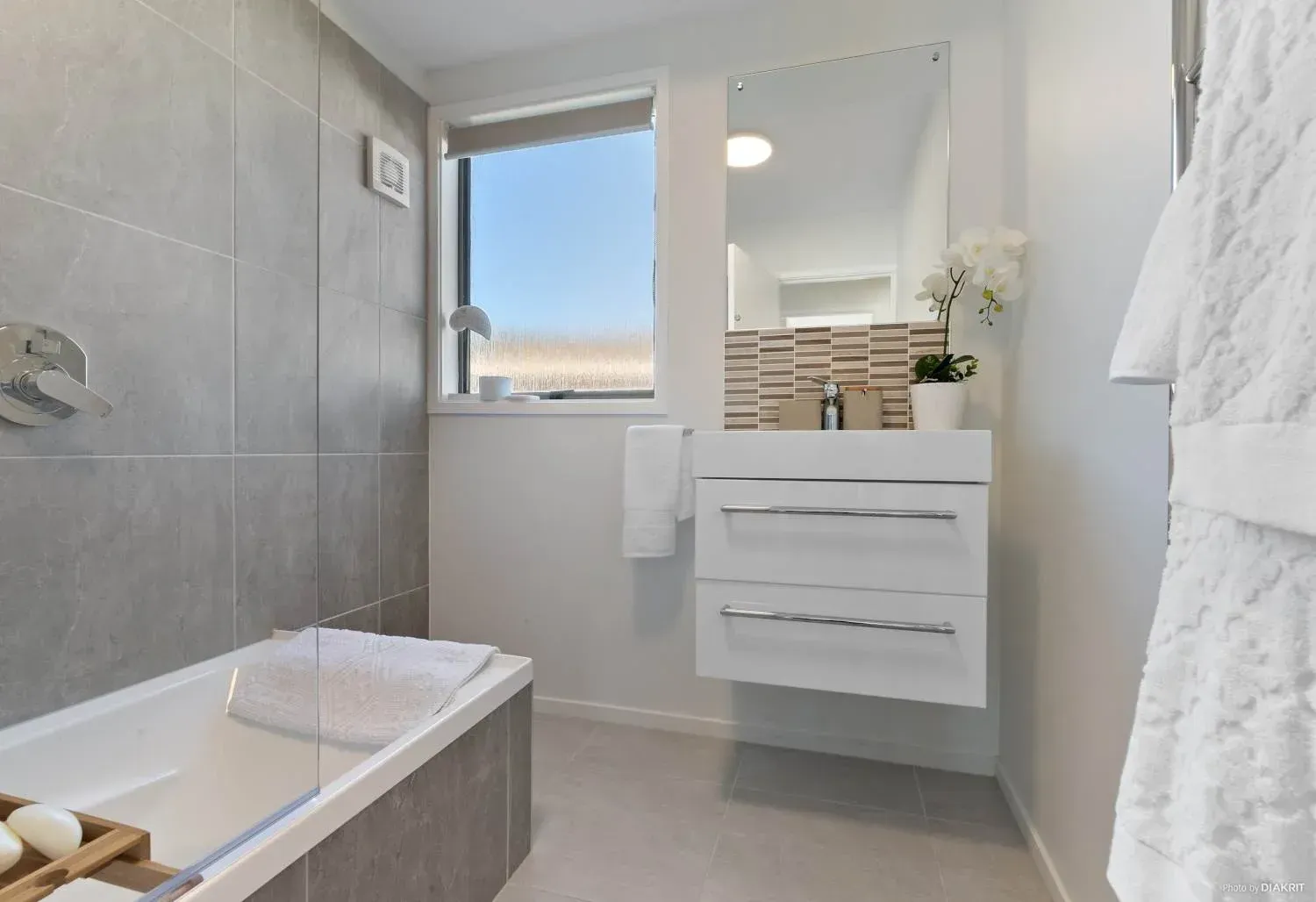 A modern bathroom featuring a white floating vanity, a gray tiled bathtub, a window, and a wall-mounted mirror.