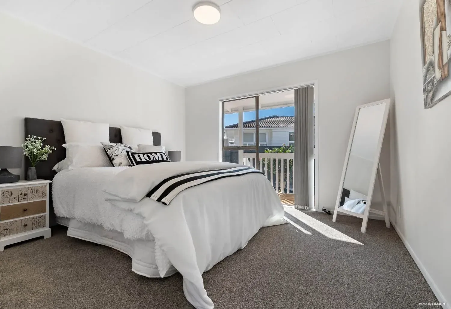 A bright bedroom featuring a queen bed with white bedding, a decorative pillow, a side table, and a full-length mirror.