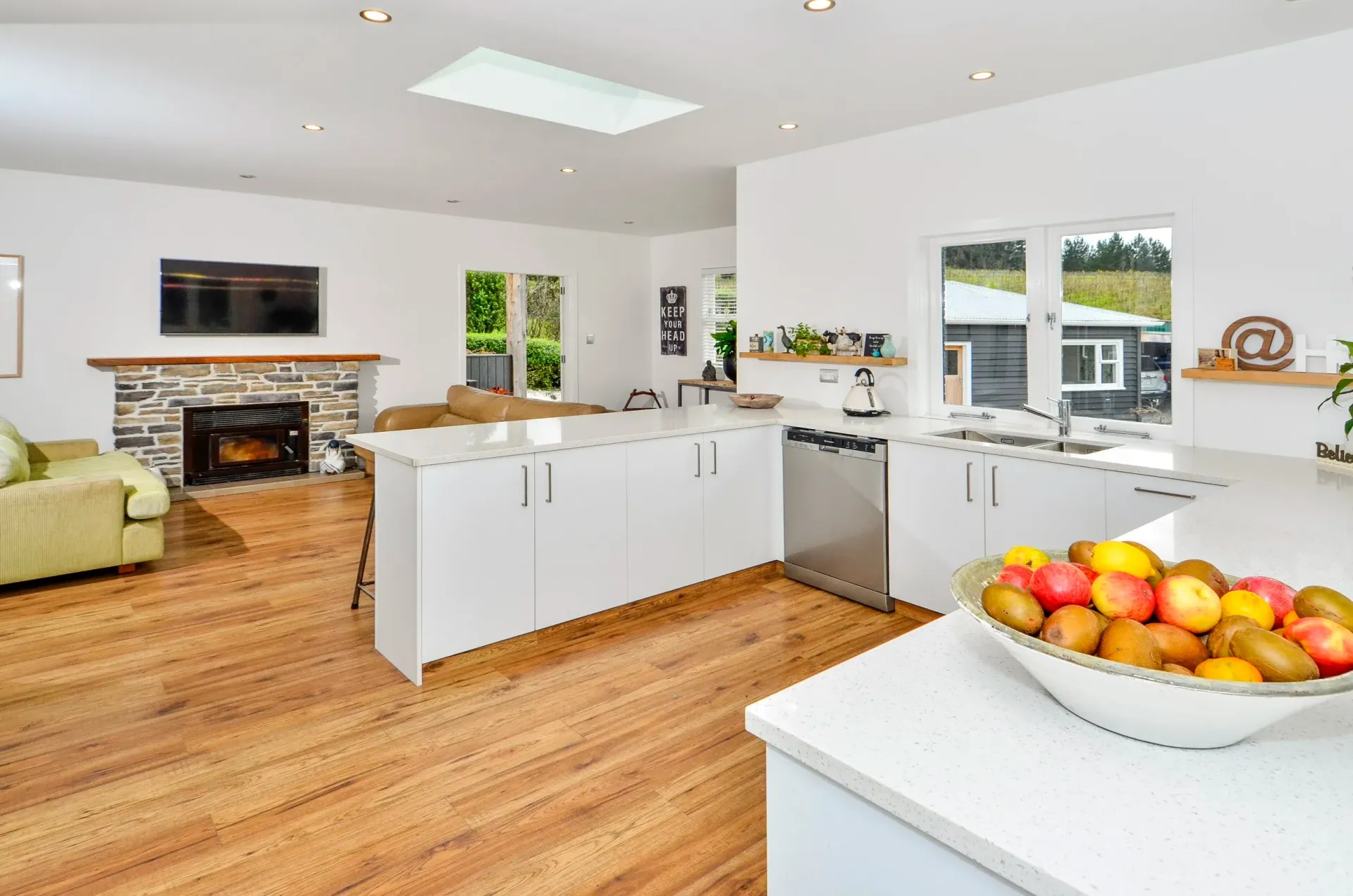 An open-plan living room and kitchen with white cabinetry, wood flooring, a fireplace, and a bowl of fruit on the counter.