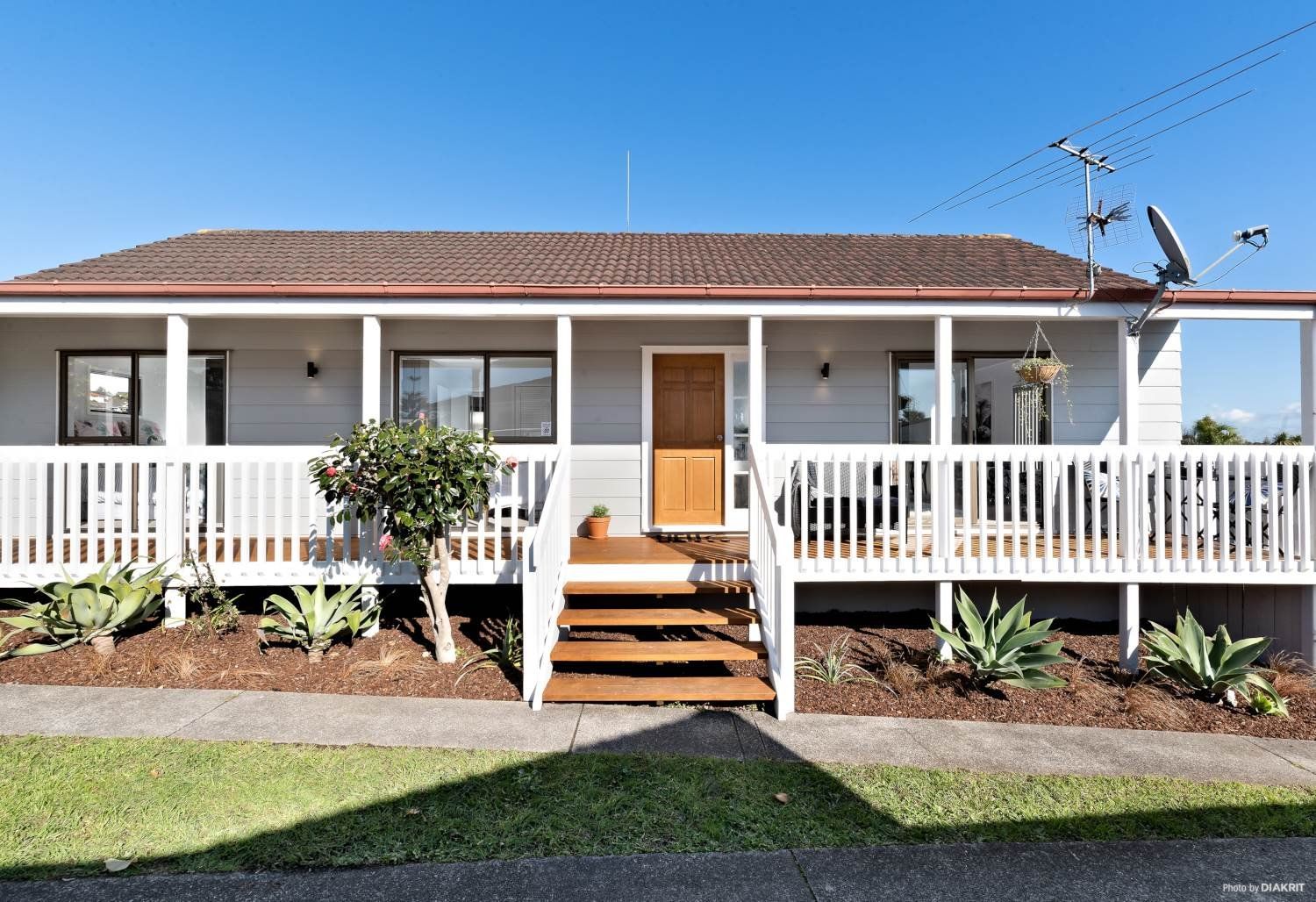 A light gray single-story house with a wooden front door, white porch railing, and landscaped yard under a blue sky.