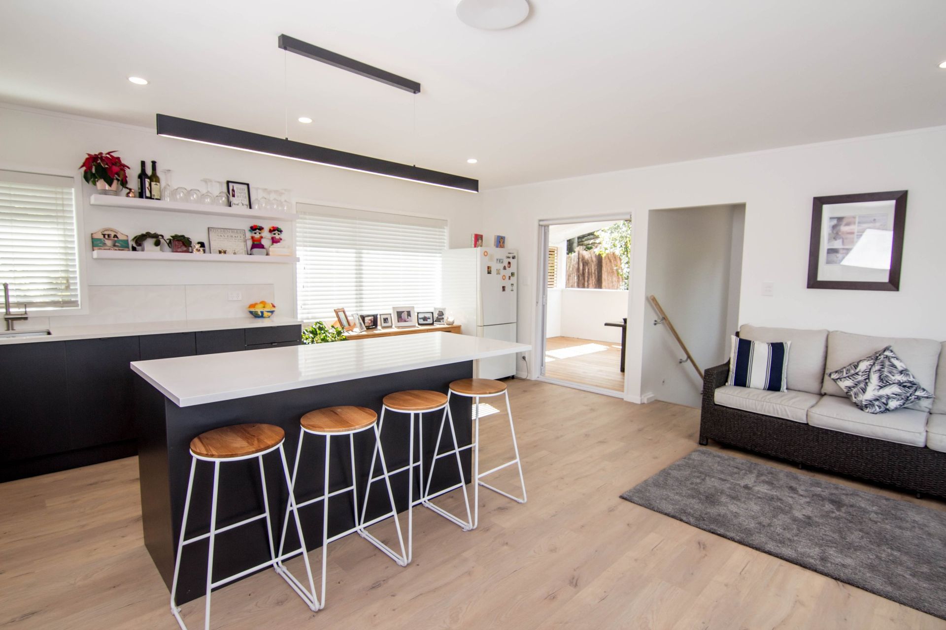 A modern, open-plan kitchen and living room featuring a dark island with four bar stools, light wood floors, and a sofa.