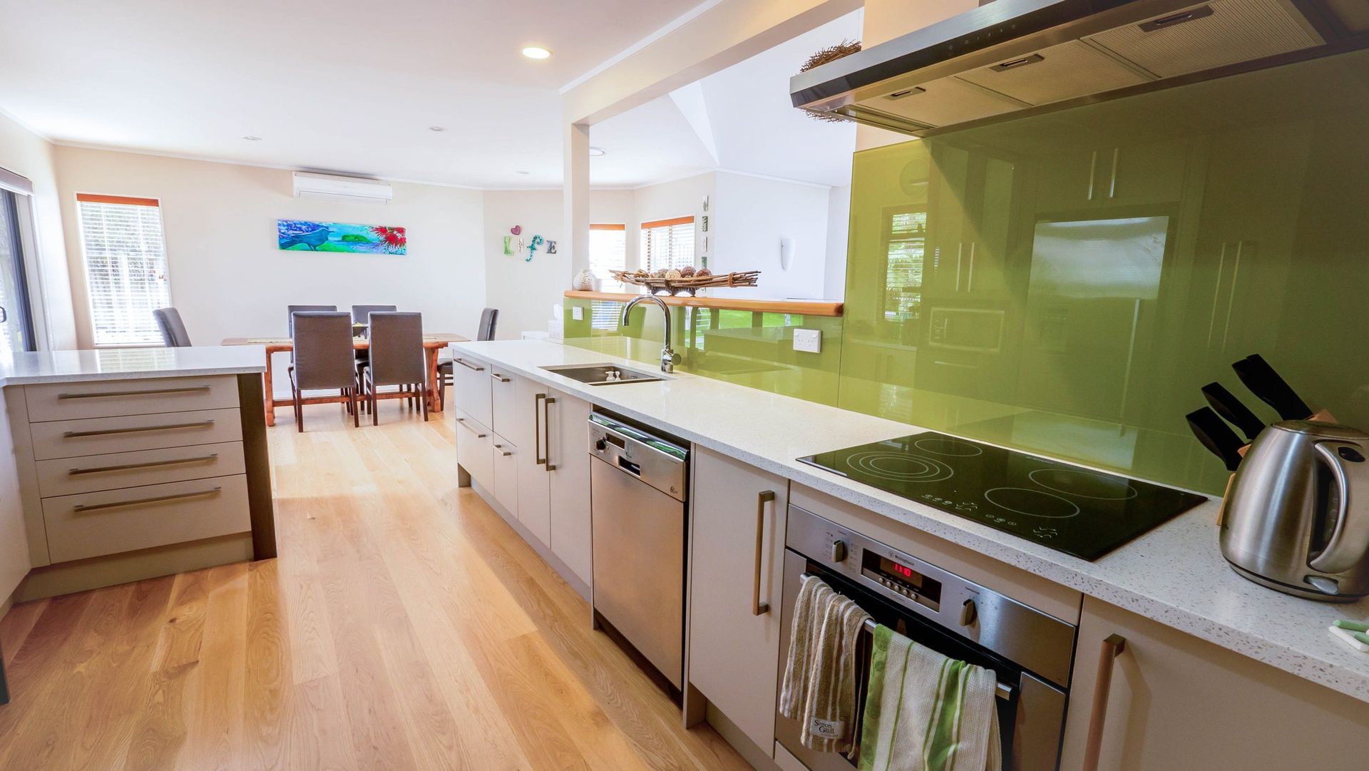 A modern, open-plan kitchen with light cabinets, a green backsplash, stainless steel appliances, and a dining area.