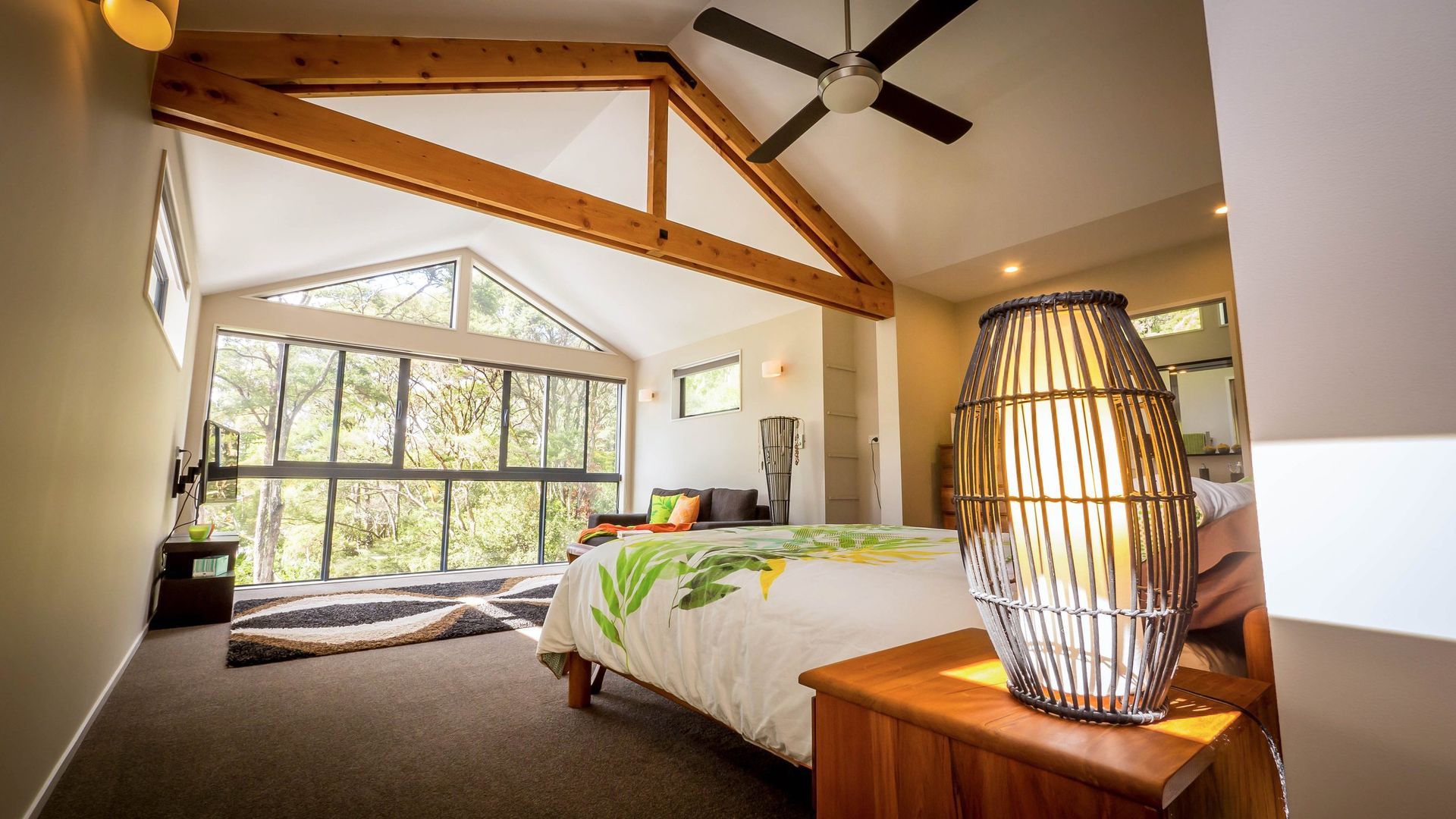 A spacious, modern bedroom with a vaulted timber-beamed ceiling, a large window wall overlooking woods, and a bed.
