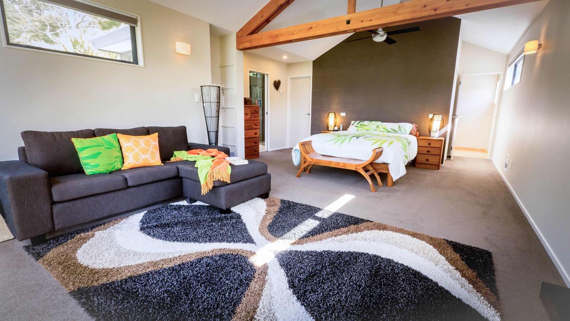 A spacious hotel room with a dark grey sofa, patterned rug, and a made bed under a high ceiling with exposed wood beams.