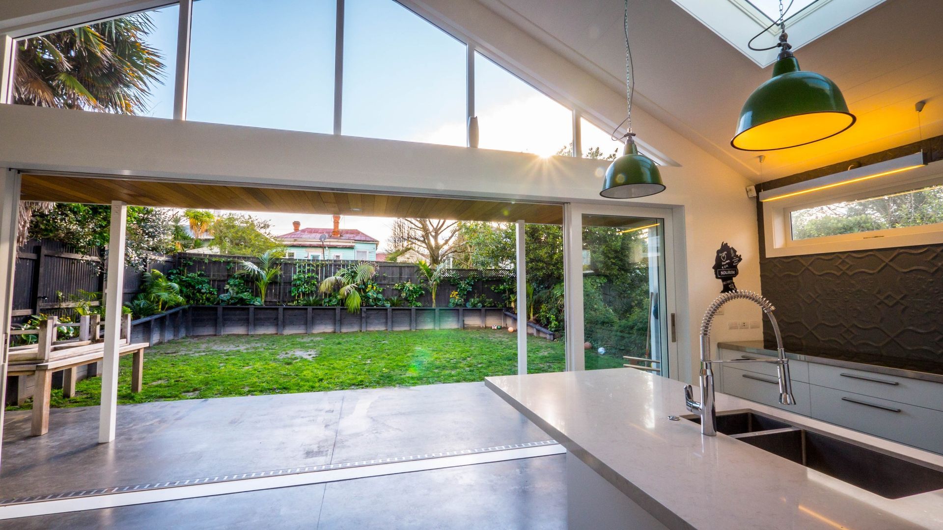 A modern open-plan kitchen with a large island, concrete floors, and floor-to-ceiling glass windows opening to a garden.