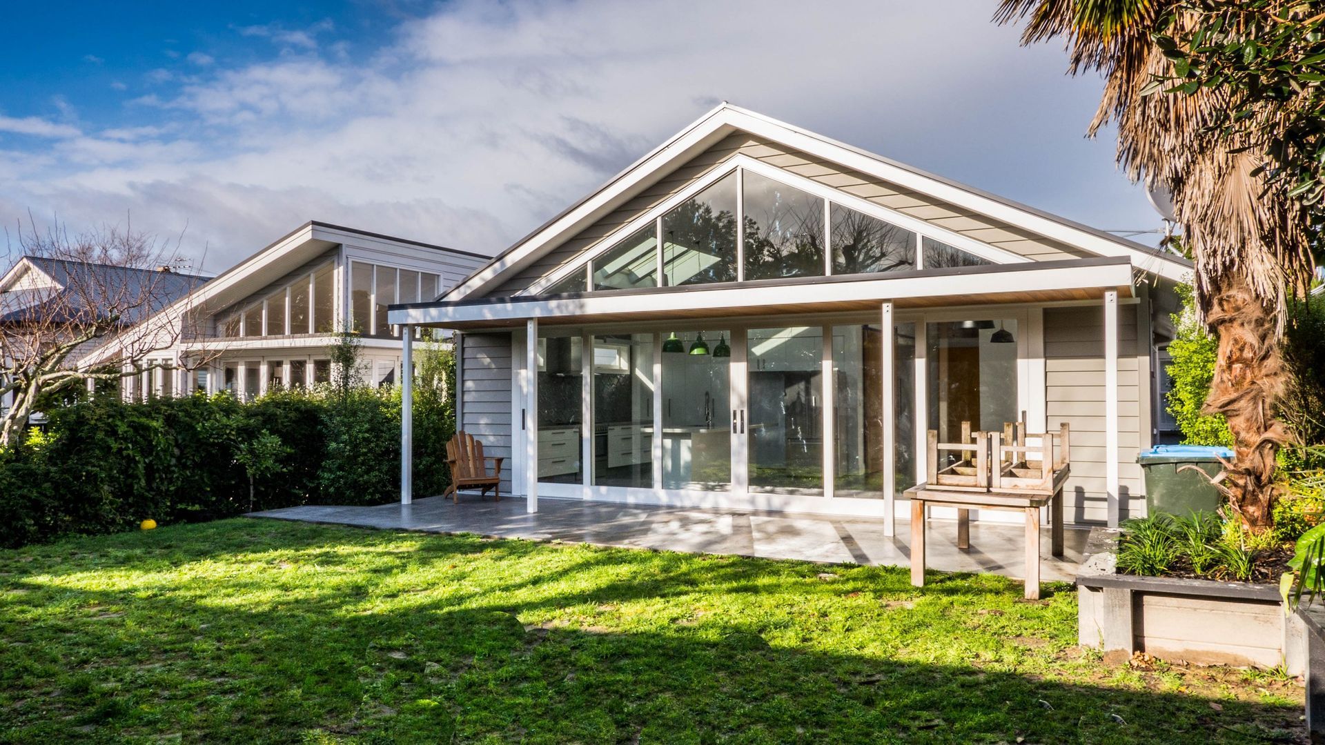 A light-colored house with large glass windows and a front patio, viewed from a sunny lawn with a palm tree on the right.