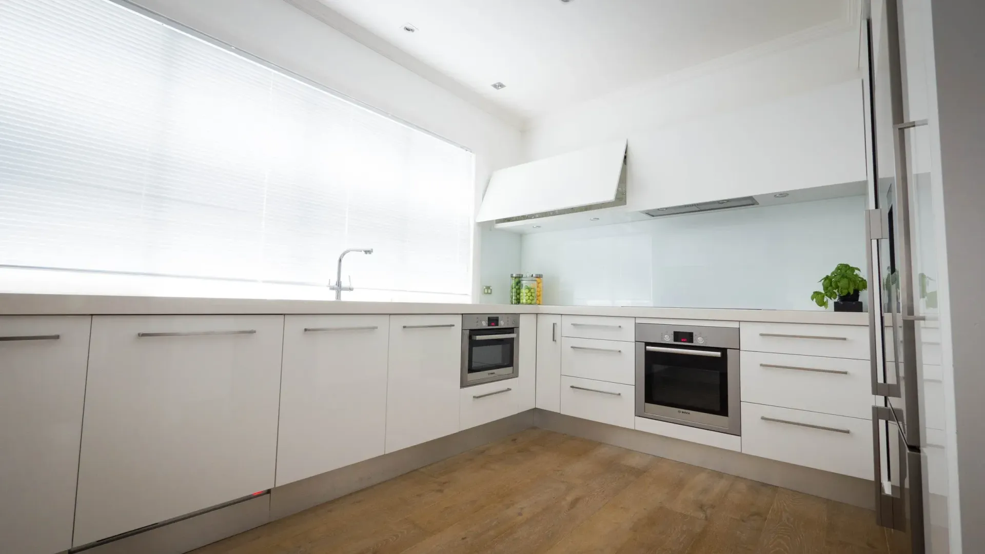 A modern, minimalist white kitchen featuring sleek cabinets, light wood floors, a built-in oven, and a large window.