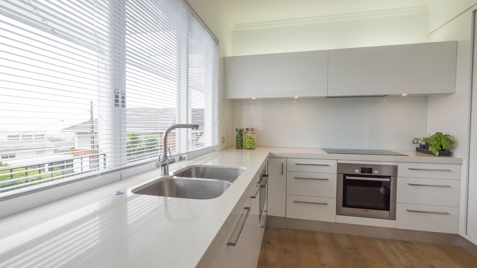 A modern kitchen with white cabinetry, a light countertop, a stainless steel sink, and large windows with white blinds.