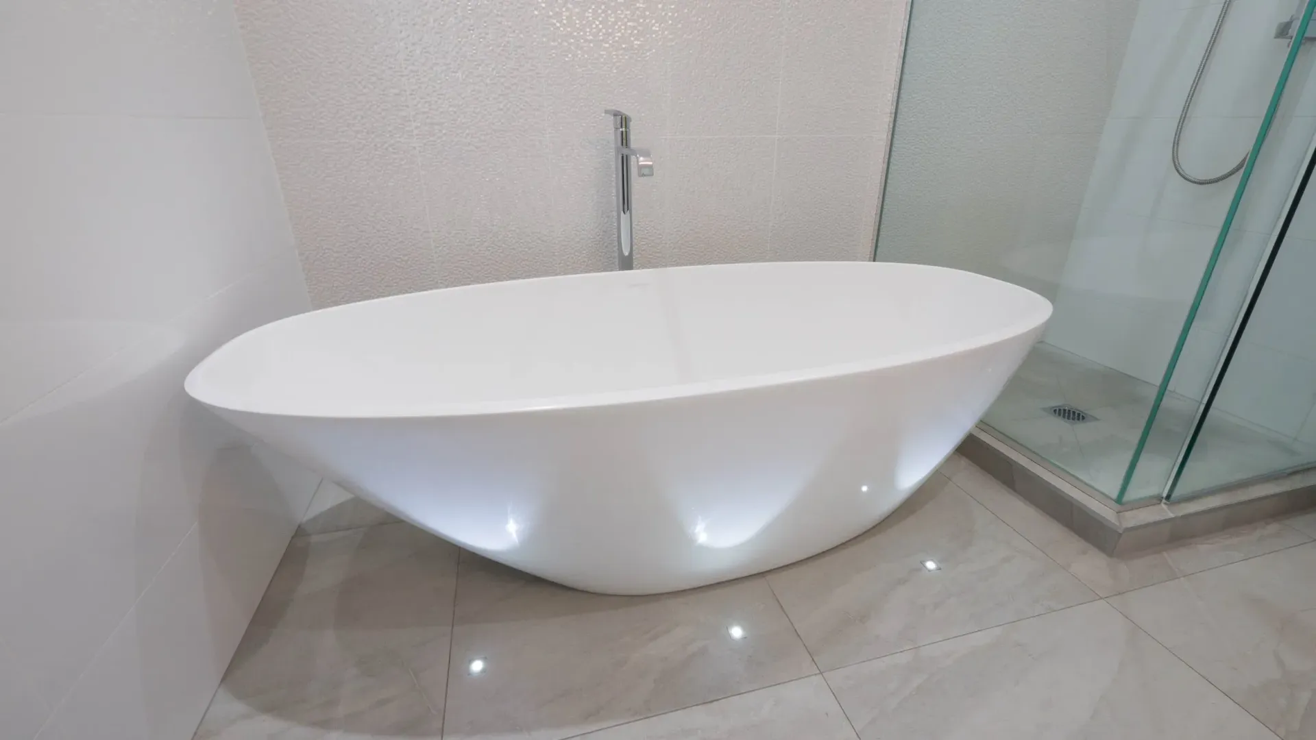A modern white oval freestanding bathtub in a tiled bathroom with a glass-enclosed shower nearby.