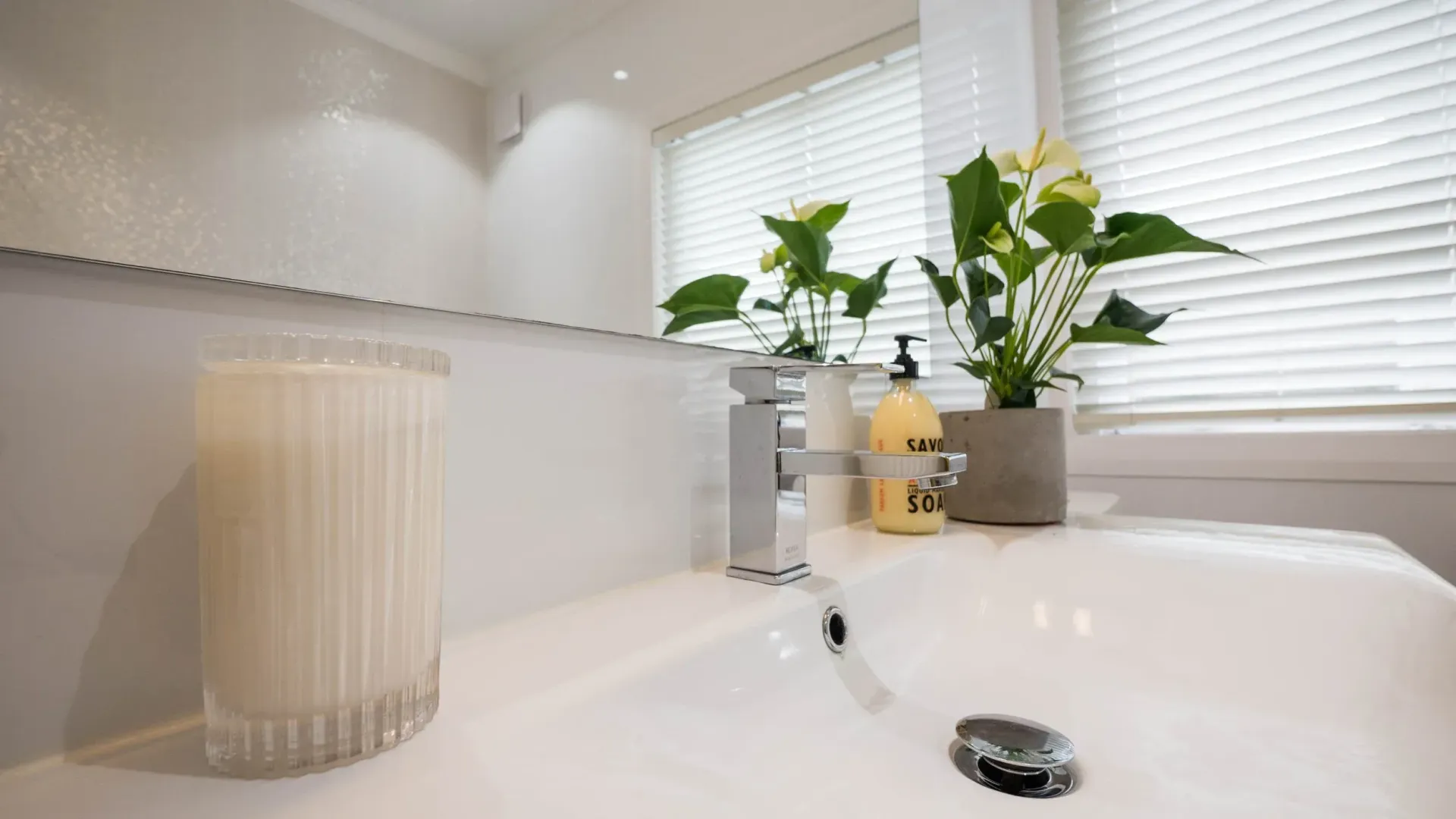 A bathroom sink with a candle, a pump soap bottle, and a potted plant in front of a mirror and blinds.