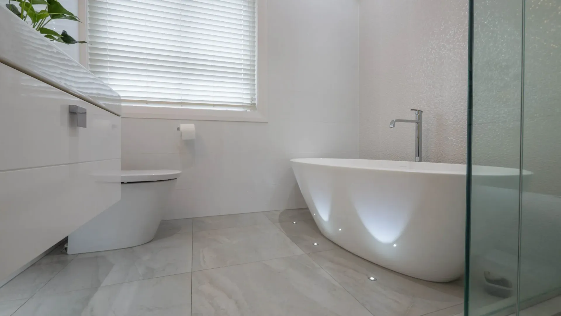 Modern bathroom with a white freestanding tub featuring under-glow lighting, a white vanity, and light-toned tiled floors.