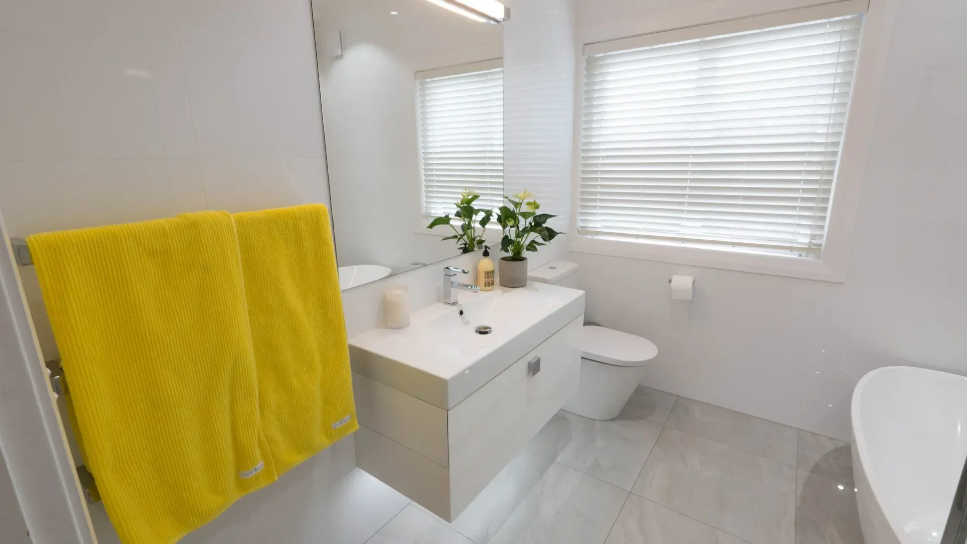 A modern bathroom with a bright yellow towel on a rack, a white vanity with plants, a toilet, and a white soaking tub.