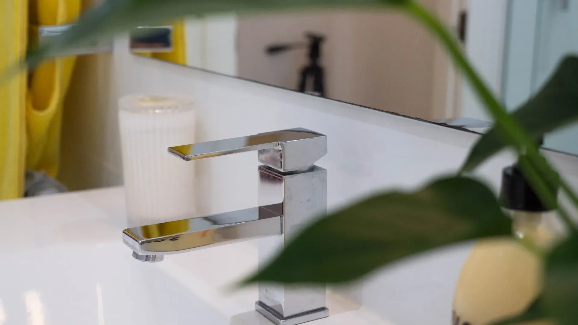 A chrome modern faucet sits on a white bathroom counter beside a white candle, partially obscured by green leaves.