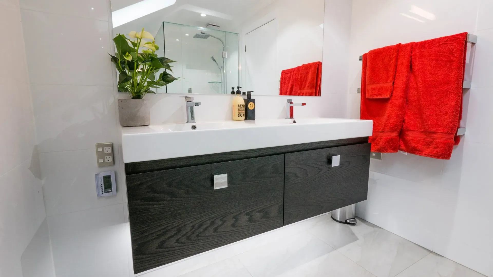 A modern bathroom with a double sink, dark textured vanity, large mirror, and bright red towels hanging on the wall.