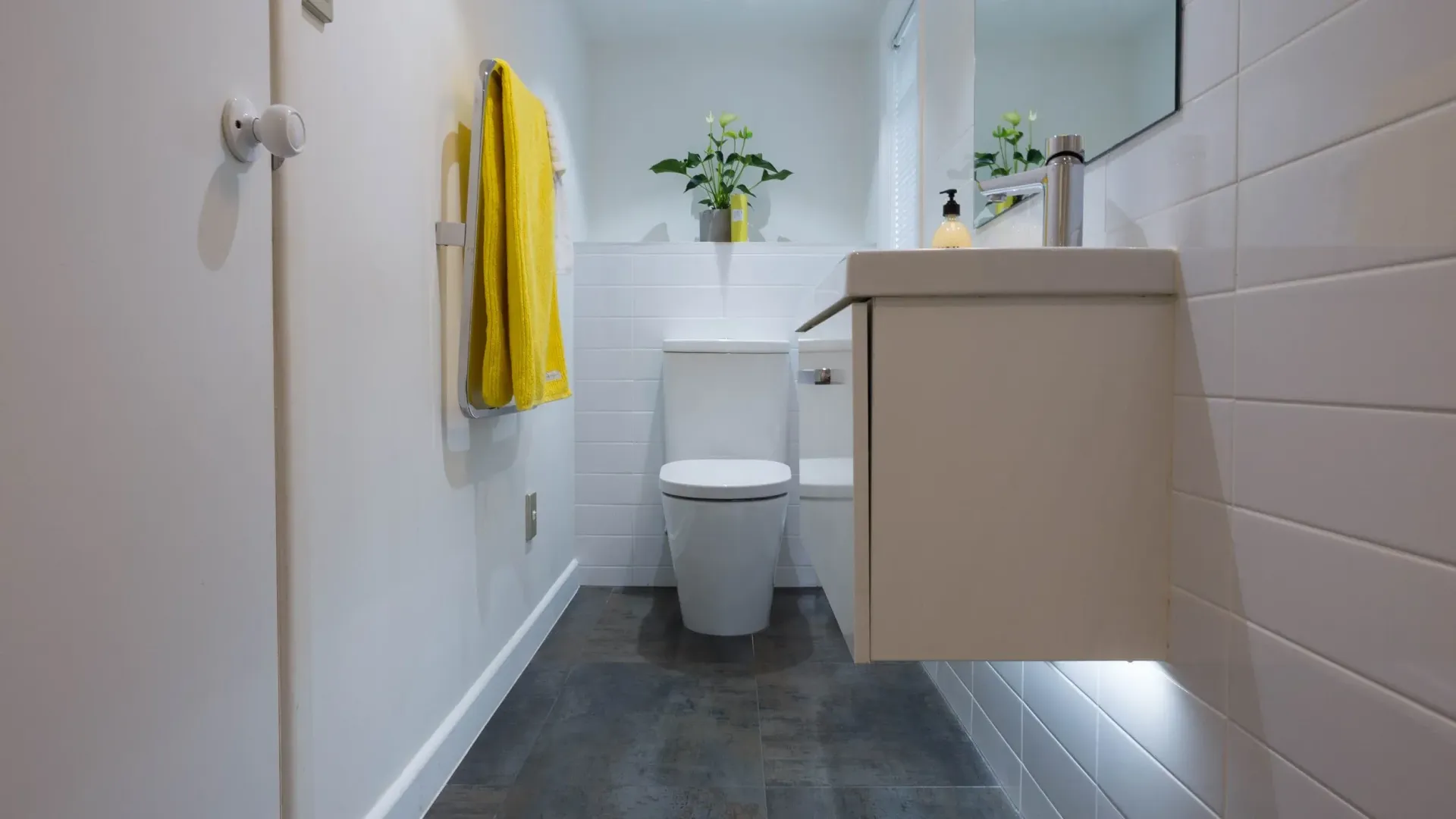 A modern bathroom features a floating vanity, a white toilet, dark tiled floors, white wall tiles, and a bright yellow towel.