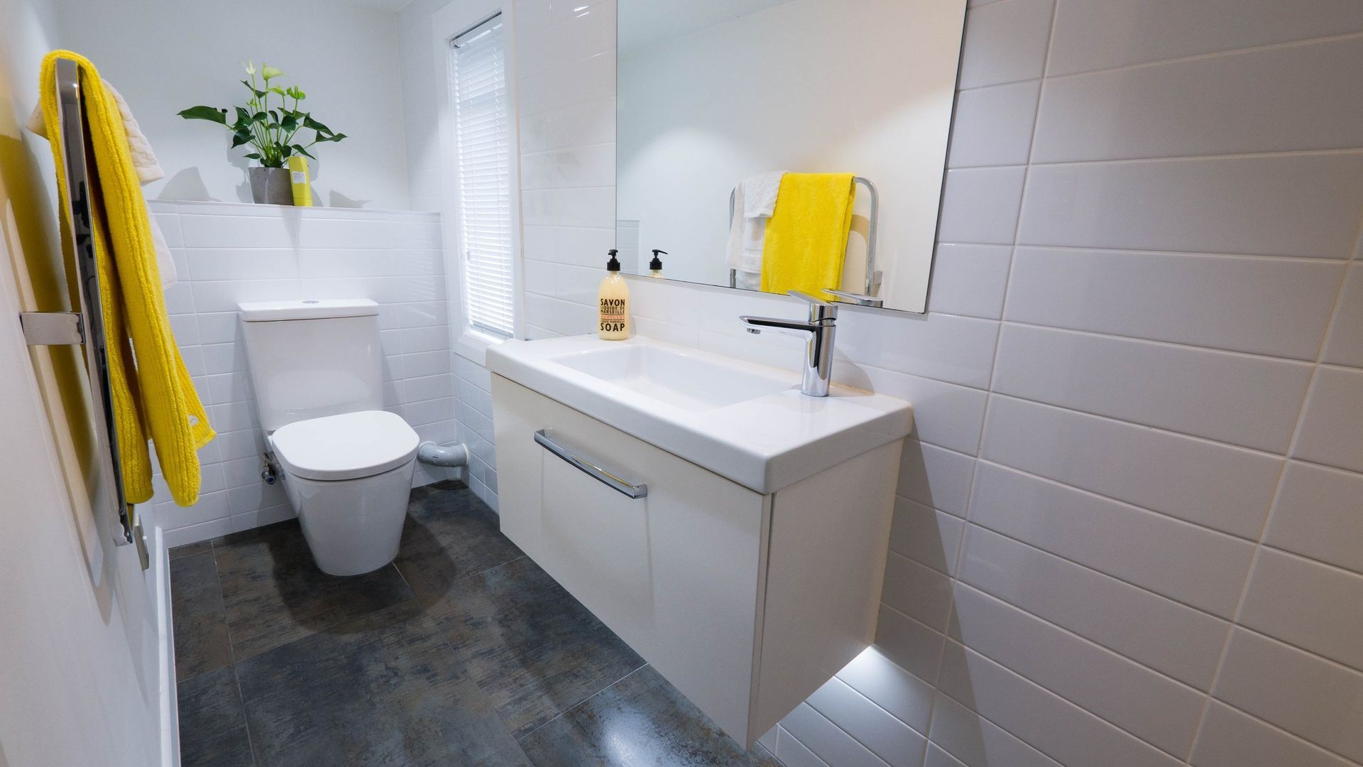 A modern white bathroom with yellow towels, a toilet, a floating vanity with a sink, and dark tiled flooring.