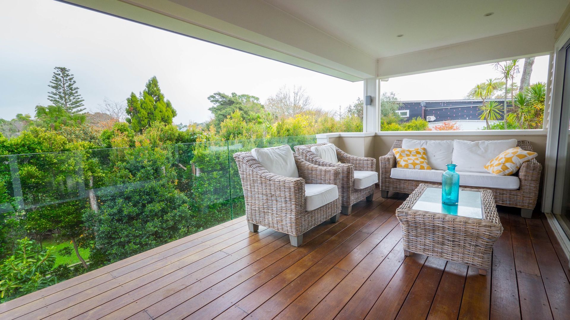 A furnished wooden deck with wicker chairs, a sofa, and a table overlooking a lush green garden.