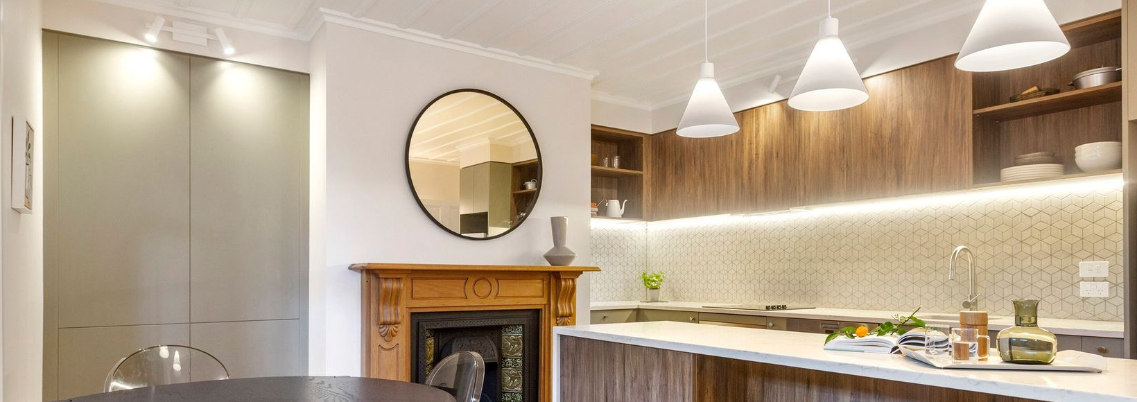 A modern kitchen with wood cabinets, a stone island, white pendant lights, and a fireplace with a round mirror above it.