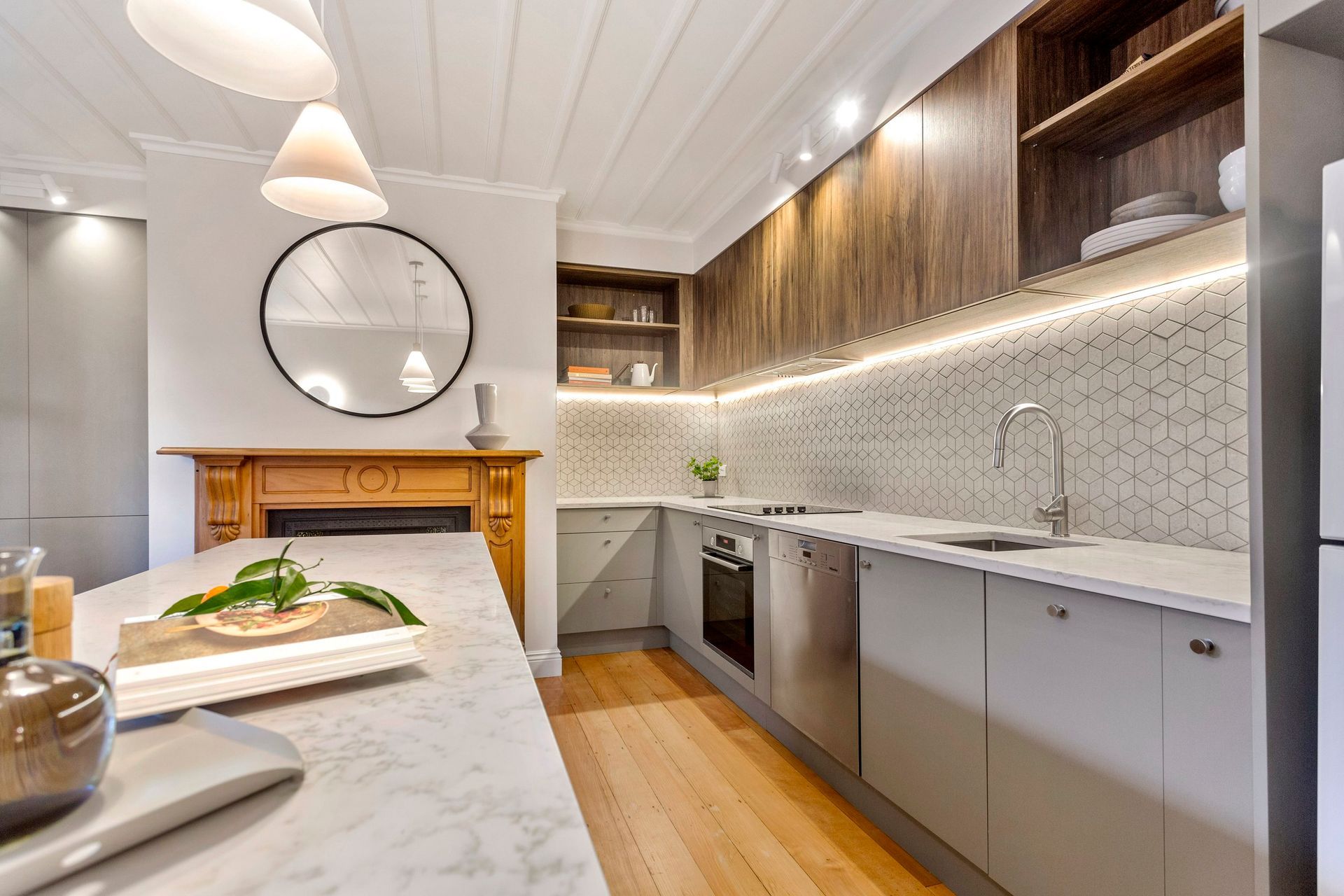 A modern kitchen with marble island, light grey cabinets, wooden upper shelves, and a patterned hexagonal tile backsplash.