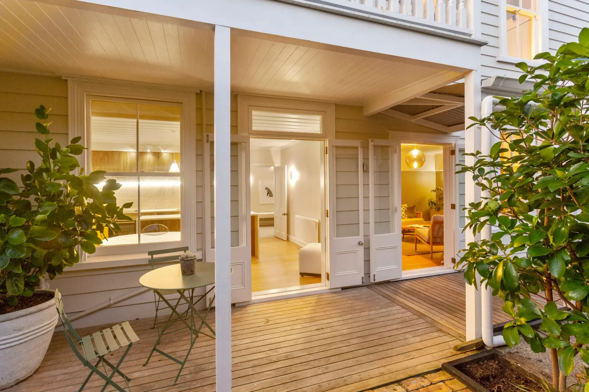 A covered wooden porch with a small bistro table and chair, looking into a brightly lit interior of a home.