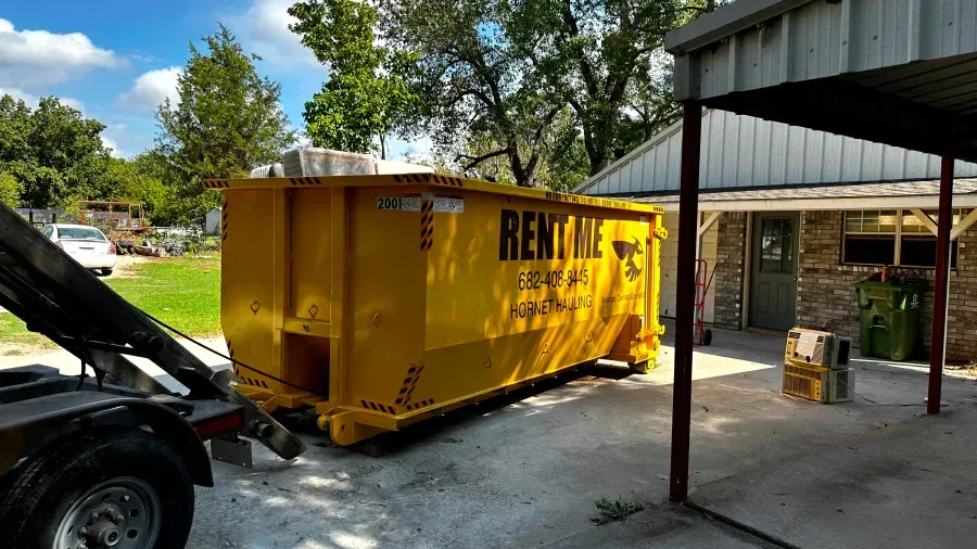 A yellow dumpster is being towed by a truck in front of a house.