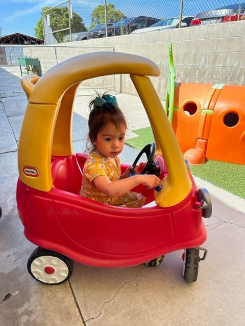 A little girl is sitting in a red and yellow toy car.