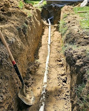A trench with a shovel and a white pipe laid in dirt, leading to a small opening at the top.