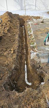 Narrow trench in muddy soil with a white pipe running vertically beside a construction site.