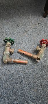 Two rusty pipe valve assemblies with green and red handles on a gray carpet, with copper pipe sections nearby