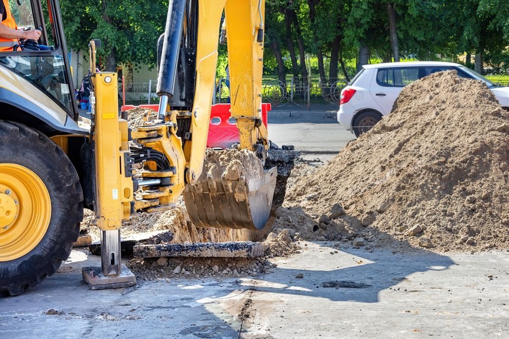 Yellow excavator digging dirt beside a road with a parked white car and soil pile in the background