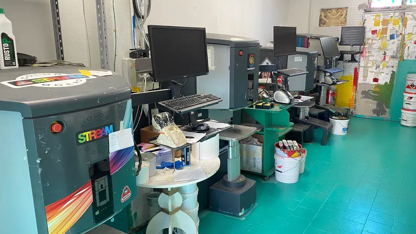 In una stanza sono disposte in fila diverse grandi macchine industriali per la colorazione dei vetri, complete di monitor e tastiere.