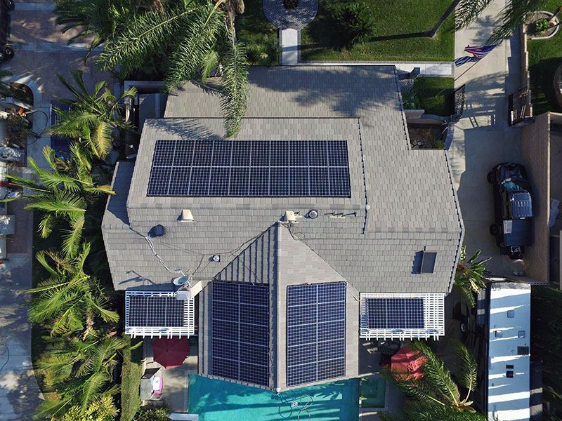 Aerial view of a house with solar panels on the roof, surrounded by trees and parked cars.